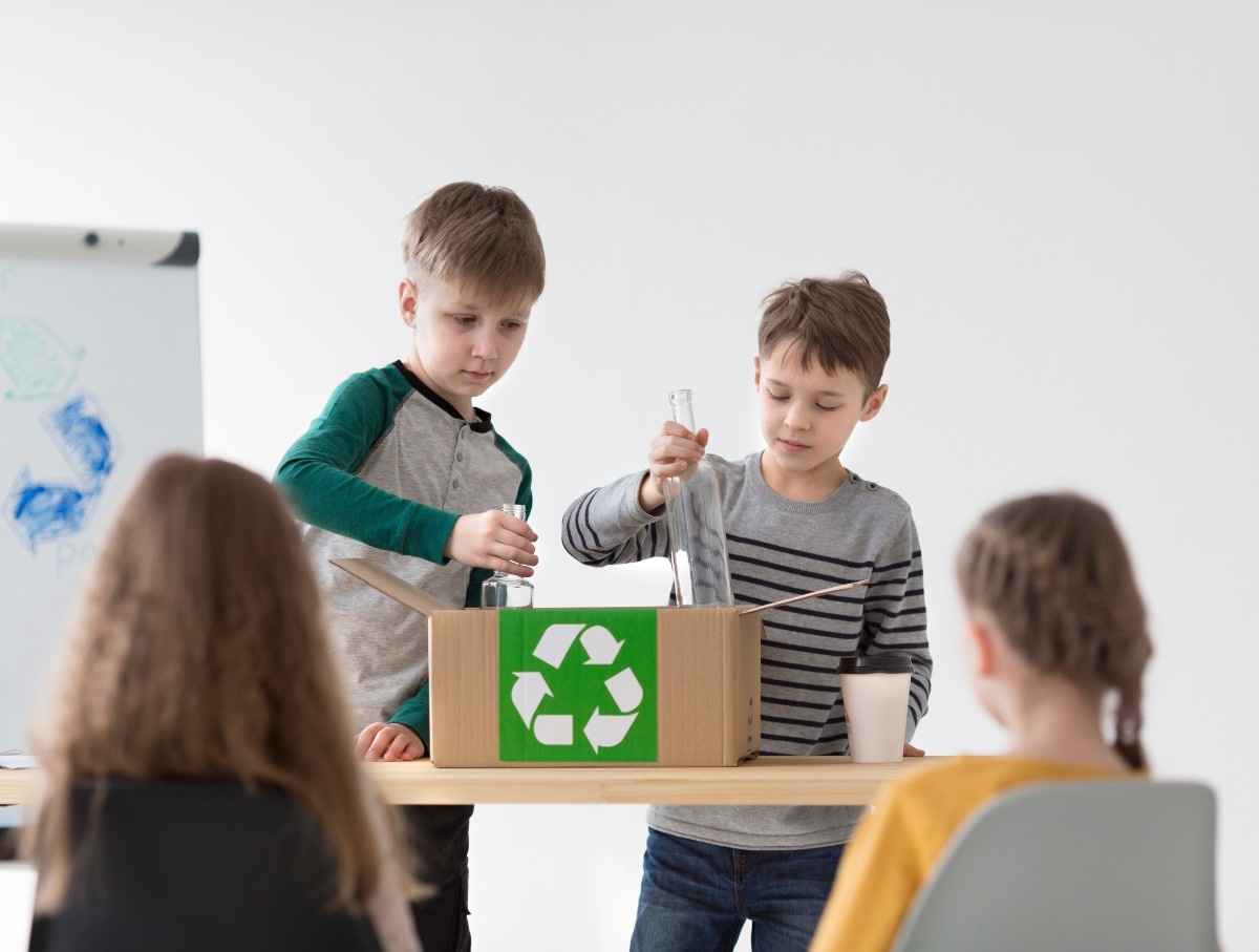 Two smiling girls holding cardboard recycling boxes standing next to a whiteboard with a colored recycling symbol and words glass, plastic, and paper.