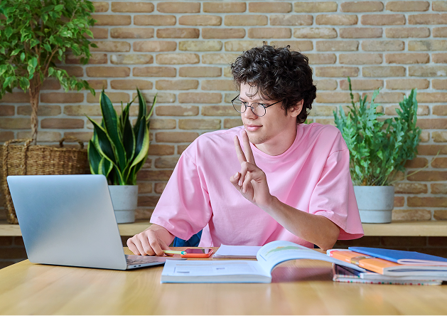 Young man in a pink shirt sitting at a table with open books and a laptop, making a peace sign with his right hand.